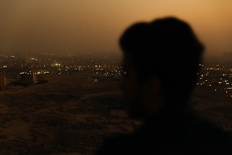 A silhouette of a person appears in the foreground overlooking a vast cityscape. The city is illuminated with an array of twinkling lights under a dusky sky, suggesting early evening or nightfall.