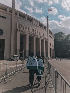 A large stadium with the words 'Estadio Municipal Paulo Machado de Carvalho' prominently displayed on its facade. In the foreground, two people wrapped in Palmeiras flags walk towards the entrance, surrounded by metal barriers. A flag waves atop a tall pole next to the building, and the sky is partly cloudy with patches of blue.