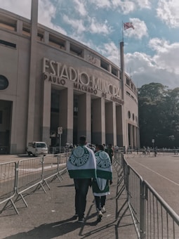 A large stadium with the words 'Estadio Municipal Paulo Machado de Carvalho' prominently displayed on its facade. In the foreground, two people wrapped in Palmeiras flags walk towards the entrance, surrounded by metal barriers. A flag waves atop a tall pole next to the building, and the sky is partly cloudy with patches of blue.