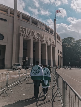 A large stadium with the words 'Estadio Municipal Paulo Machado de Carvalho' prominently displayed on its facade. In the foreground, two people wrapped in Palmeiras flags walk towards the entrance, surrounded by metal barriers. A flag waves atop a tall pole next to the building, and the sky is partly cloudy with patches of blue.