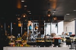 A stylish and modern restaurant interior with decorative hanging lamps, a variety of colorful fruits displayed in baskets, and a slicing machine on the counter. The background includes a bar area with bottles and glasses, as well as tables and chairs set for dining.