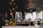 A stylish and modern restaurant interior with decorative hanging lamps, a variety of colorful fruits displayed in baskets, and a slicing machine on the counter. The background includes a bar area with bottles and glasses, as well as tables and chairs set for dining.