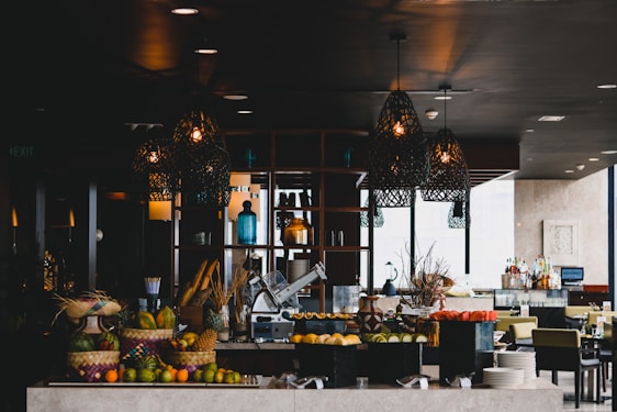 A stylish and modern restaurant interior with decorative hanging lamps, a variety of colorful fruits displayed in baskets, and a slicing machine on the counter. The background includes a bar area with bottles and glasses, as well as tables and chairs set for dining.