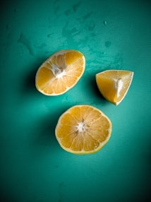 Three pieces of a sliced lemon are arranged on a textured green background. The lemon segments are positioned with two halves and one quarter piece, displaying the juicy and vibrant interior. There are small droplets of water around the segments, adding a fresh and appetizing look.