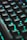 A close-up view of a computer keyboard with keys illuminated by vibrant LED backlighting, predominantly in shades of teal and green. The keys are prominently featured, creating a futuristic and tech-focused atmosphere.