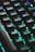 A close-up view of a computer keyboard with keys illuminated by vibrant LED backlighting, predominantly in shades of teal and green. The keys are prominently featured, creating a futuristic and tech-focused atmosphere.