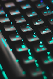 A close-up view of a computer keyboard with keys illuminated by vibrant LED backlighting, predominantly in shades of teal and green. The keys are prominently featured, creating a futuristic and tech-focused atmosphere.
