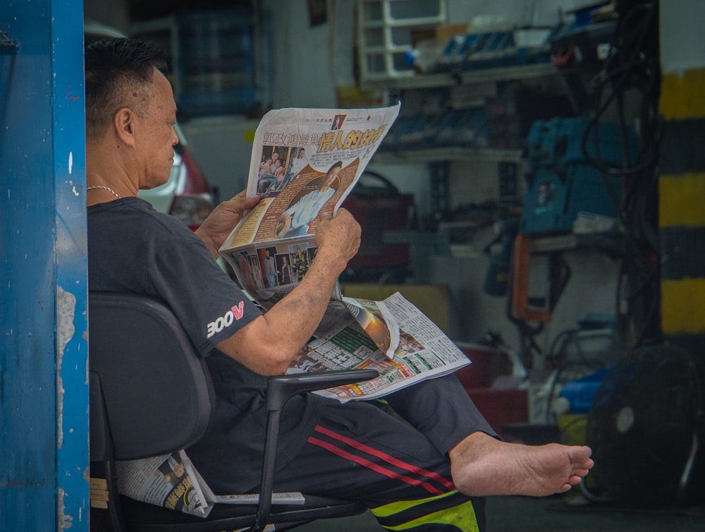 man wearing black shirt sitting on chair reading newspaper