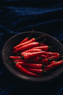 A dark, moody photograph featuring a black bowl filled with vibrant red chili peppers. The background is a deep blue fabric with subtle checkered patterns, enhancing the rich colors of the peppers.