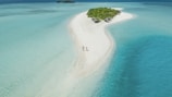 Aerial view of a long, narrow sandbar surrounded by clear, turquoise waters of the ocean. The sandbar leads to a small, lush green island with dense vegetation and a few thatched-roof huts. Two people are walking hand-in-hand along the sandy path, enjoying the serene and picturesque setting.