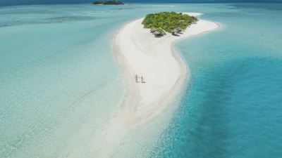 Aerial view of a long, narrow sandbar surrounded by clear, turquoise waters of the ocean. The sandbar leads to a small, lush green island with dense vegetation and a few thatched-roof huts. Two people are walking hand-in-hand along the sandy path, enjoying the serene and picturesque setting.