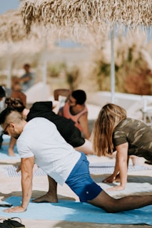 Several people are practicing yoga outdoors on a sunny day, performing a stretching pose on blue mats. They are under straw sun umbrellas, suggesting a beach or outdoor setting. The background is slightly blurred with hints of greenery.