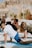 Several people are practicing yoga outdoors on a sunny day, performing a stretching pose on blue mats. They are under straw sun umbrellas, suggesting a beach or outdoor setting. The background is slightly blurred with hints of greenery.