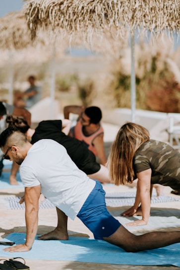 Several people are practicing yoga outdoors on a sunny day, performing a stretching pose on blue mats. They are under straw sun umbrellas, suggesting a beach or outdoor setting. The background is slightly blurred with hints of greenery.