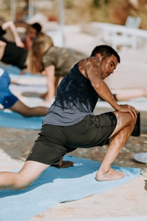 A person is engaging in a yoga or stretching exercise outdoors on a sunny day, positioned on a light blue mat. Others can be seen in the background also performing similar activities. The scene captures a sense of focus and balance.