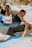 A person is engaging in a yoga or stretching exercise outdoors on a sunny day, positioned on a light blue mat. Others can be seen in the background also performing similar activities. The scene captures a sense of focus and balance.