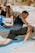 A person is engaging in a yoga or stretching exercise outdoors on a sunny day, positioned on a light blue mat. Others can be seen in the background also performing similar activities. The scene captures a sense of focus and balance.