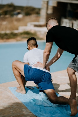 Two people are by a poolside; one is kneeling on a towel while another supports him by holding his lower back. The scene is relaxed and sunny, with a focus on physical guidance or exercise.