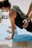 A person is in a yoga or stretching pose on a blue mat, outdoors. Another person's hands are assisting or providing support on the back. The setting appears sunny, with shadows cast on the ground.