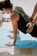 A person is in a yoga or stretching pose on a blue mat, outdoors. Another person's hands are assisting or providing support on the back. The setting appears sunny, with shadows cast on the ground.