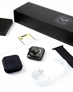 A smartwatch is displayed with its accessories, including a black watch strap, a white charging cable, and packaging materials. Everything is neatly arranged on a white surface, emphasizing the sleek design of the watch and its components.