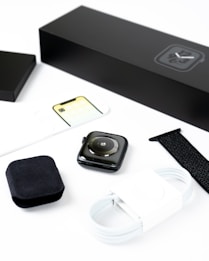 A smartwatch is displayed with its accessories, including a black watch strap, a white charging cable, and packaging materials. Everything is neatly arranged on a white surface, emphasizing the sleek design of the watch and its components.