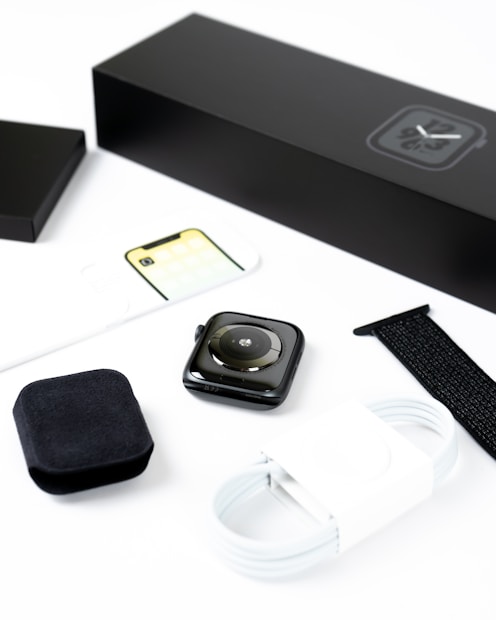 A smartwatch is displayed with its accessories, including a black watch strap, a white charging cable, and packaging materials. Everything is neatly arranged on a white surface, emphasizing the sleek design of the watch and its components.