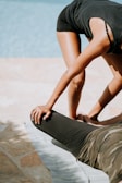 Two people are engaging in an outdoor stretching session by a pool. One individual is bending forward while supporting the leg of another person lying on a mat. The stretching exercise seems to focus on the legs, with the standing person extending support using hands placed on the other person's thigh.