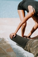 Two people are engaging in an outdoor stretching session by a pool. One individual is bending forward while supporting the leg of another person lying on a mat. The stretching exercise seems to focus on the legs, with the standing person extending support using hands placed on the other person's thigh.