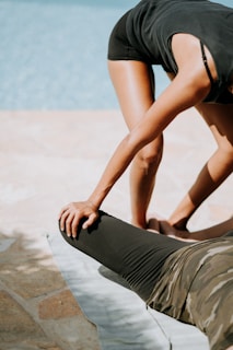 Two people are engaging in an outdoor stretching session by a pool. One individual is bending forward while supporting the leg of another person lying on a mat. The stretching exercise seems to focus on the legs, with the standing person extending support using hands placed on the other person's thigh.