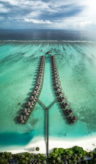An aerial view of a luxurious resort featuring overwater bungalows arranged in two parallel rows with a connecting walkway over a clear, turquoise ocean. The calm sea stretches towards the horizon where it meets the sky, filled with clouds. The foreground shows a pristine sandy beach and lush green palm trees.