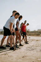 A group of five people, dressed in athletic wear, are gathered on a sandy beach. They appear to be preparing for an activity or exercise, focusing intently on their posture. The background is slightly blurred, suggesting an outdoor setting with a clear sky.