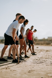 A group of five people, dressed in athletic wear, are gathered on a sandy beach. They appear to be preparing for an activity or exercise, focusing intently on their posture. The background is slightly blurred, suggesting an outdoor setting with a clear sky.
