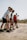 A group of five people, dressed in athletic wear, are gathered on a sandy beach. They appear to be preparing for an activity or exercise, focusing intently on their posture. The background is slightly blurred, suggesting an outdoor setting with a clear sky.