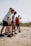 A group of five people, dressed in athletic wear, are gathered on a sandy beach. They appear to be preparing for an activity or exercise, focusing intently on their posture. The background is slightly blurred, suggesting an outdoor setting with a clear sky.