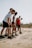 A group of five people, dressed in athletic wear, are gathered on a sandy beach. They appear to be preparing for an activity or exercise, focusing intently on their posture. The background is slightly blurred, suggesting an outdoor setting with a clear sky.