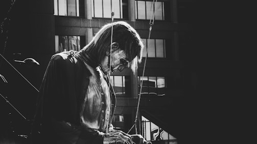 A black and white image featuring a large puppet or mannequin in an urban setting. The puppet appears detailed and lifelike, with a rope or mechanism for controlling its movements visible. It is positioned in front of a building with numerous windows, casting dramatic shadows.