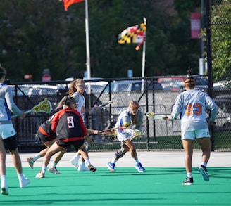 A group of athletes engage in a lacrosse game on a field with a green surface. They are wearing sports gear, including protective eyewear and jerseys with numbers. A flag in the background waves above a chain-link fence, and there are trees and parked cars visible beyond the fence.