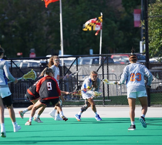A group of athletes engage in a lacrosse game on a field with a green surface. They are wearing sports gear, including protective eyewear and jerseys with numbers. A flag in the background waves above a chain-link fence, and there are trees and parked cars visible beyond the fence.
