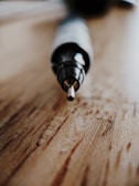A close-up view of a pen resting on a wooden surface, focused on the pen tip. The background is softly blurred, giving prominence to the pen's metallic tip and sleek design.