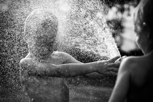 Two children are playing joyfully with water on a sunny day. The spray of water droplets is captured in motion, creating a refreshing and playful scene. The image is in black and white, emphasizing the texture of the water and the expressions of the children.