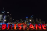 Large illuminated letters spell out 'BRISBANE' in vibrant red, set against a backdrop of a modern city skyline at night. Tall buildings are adorned with colorful lights, contributing to the lively urban atmosphere.