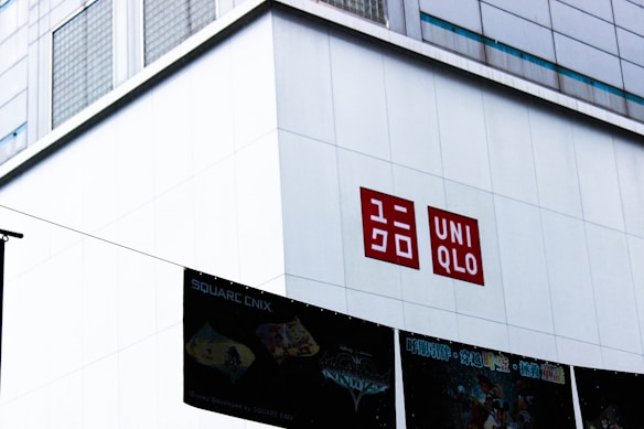 A large building facade with a prominent red and white UNIQLO logo displayed on its wall. Below the logo are several banners with brand names and imagery, suggesting a commercial area.