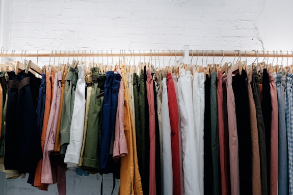 A row of various colored pants hanging on wooden hangers against a white brick wall. The garments include different shades of green, blue, red, pink, orange, and beige, neatly arranged on a rack.