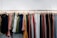 A row of various colored pants hanging on wooden hangers against a white brick wall. The garments include different shades of green, blue, red, pink, orange, and beige, neatly arranged on a rack.