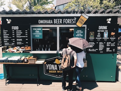 A food and drink stall named 'Omohara Beer Forest' with a menu offering a wide selection of craft beers and food options. Two people are standing in front of the stall, one holding an umbrella. The setup has a cozy, casual outdoor atmosphere with greenery in the background.