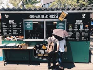 A food and drink stall named 'Omohara Beer Forest' with a menu offering a wide selection of craft beers and food options. Two people are standing in front of the stall, one holding an umbrella. The setup has a cozy, casual outdoor atmosphere with greenery in the background.