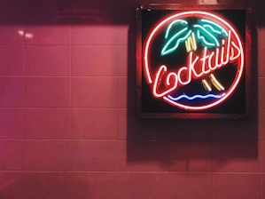 A neon sign features the word 'Cocktails' with palm trees and waves in bright colors against a pink tiled wall.