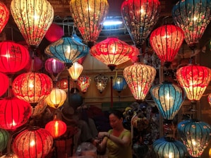 A vibrant display of colorful, intricately designed lanterns hanging from the ceiling, predominantly in shades of red and blue. The lanterns are illuminated, casting a warm glow that enhances their floral and ornate patterns. Below them, a person is engaged in handling or organizing something, surrounded by the soft lighting.