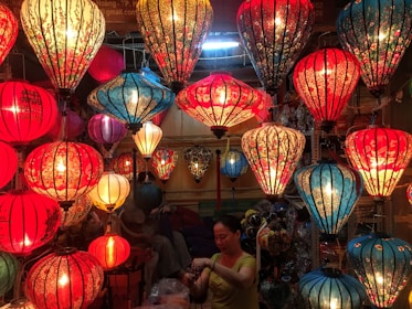 A vibrant display of colorful, intricately designed lanterns hanging from the ceiling, predominantly in shades of red and blue. The lanterns are illuminated, casting a warm glow that enhances their floral and ornate patterns. Below them, a person is engaged in handling or organizing something, surrounded by the soft lighting.